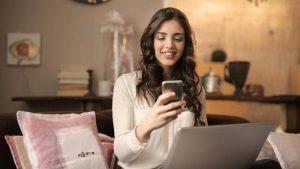 A smiling woman with long brown hair uses a smartphone while sitting on a sofa with a laptop. The cozy room feels warm and inviting, with soft lighting.