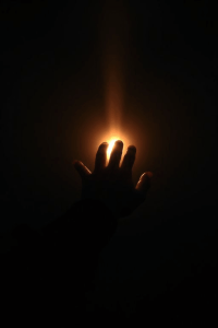 A silhouetted hand reaches towards a warm, glowing light in darkness, with fingers spread wide, conveying mystery and a search for illumination.