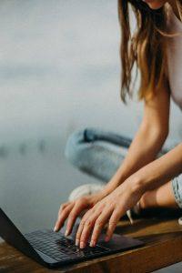 A woman in casual jeans types on a laptop outdoors, sitting cross-legged on a wooden platform. The setting is relaxed, with soft, natural lighting.