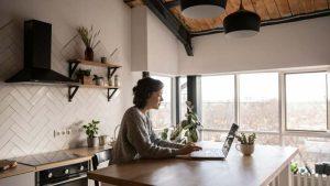 A person in a cozy kitchen is focused on typing on a laptop at a wooden table. Sunlight streams through large windows, illuminating plants and kitchen decor.