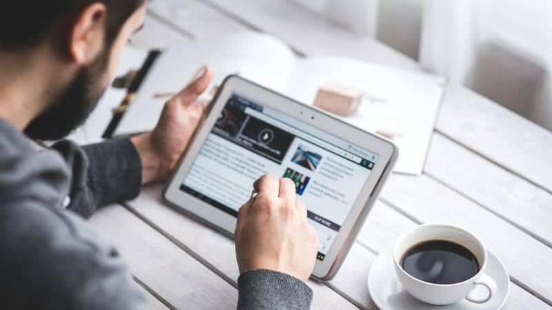 A man sits at a table, using a tablet to browse online news, with a cup of coffee nearby. The setting is casual and focused on digital media.