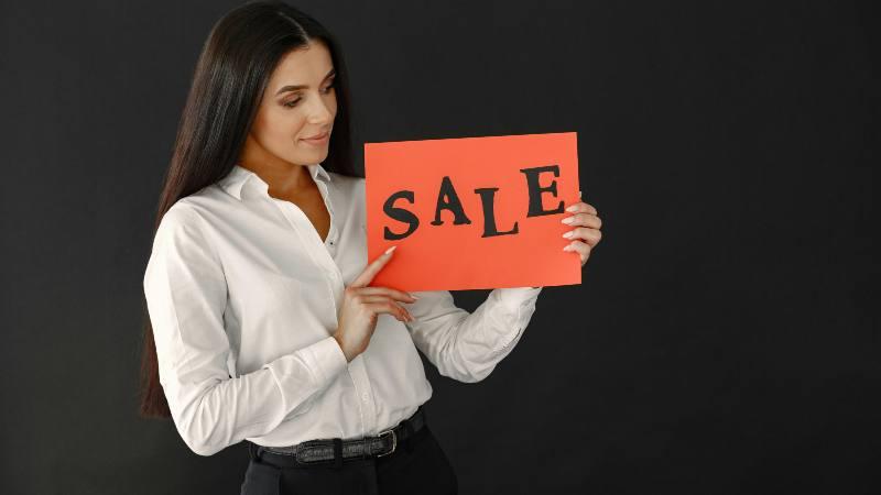 A woman in a white blouse holds a red sign with "SALE" written in bold black letters against a plain black background, conveying a promotional tone.