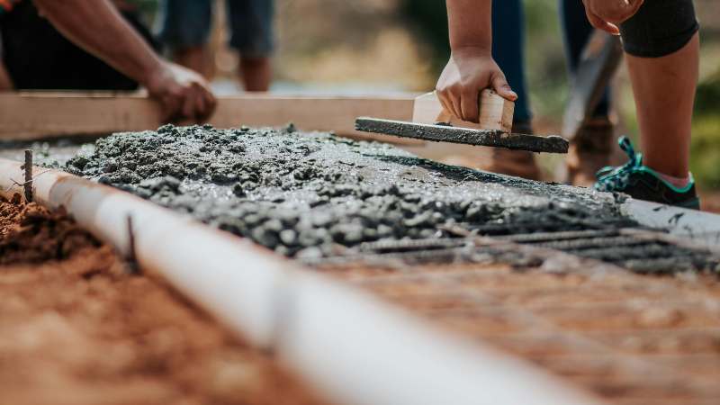 Construction workers smoothing wet concrete with tools on a sunny day. The focus is on hands and equipment, conveying teamwork and manual labor.
