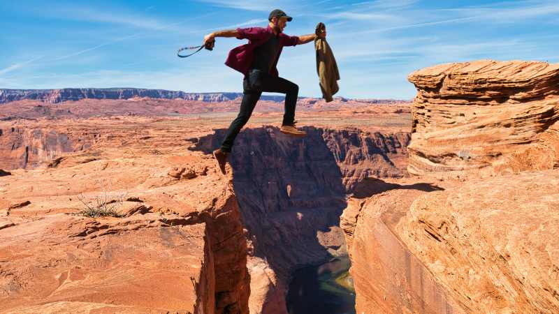 A person wearing a cap and holding a jacket leaps across a vast canyon with red rock formations under a clear blue sky, conveying adventure and daring.